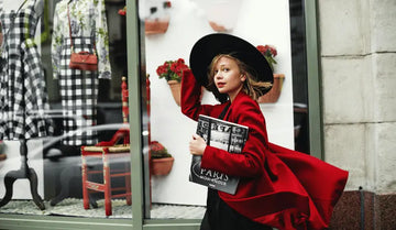 Woman in a red coat and black hat holding a magazine in front of a store window display.
