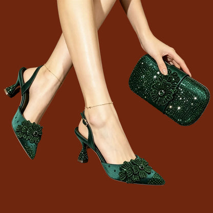A pair of dark green pointed-toe slingback heels with large floral embellishments and high stiletto heels, shown on a model's feet. The model is also holding a matching dark green rhinestone-embellished clutch bag.