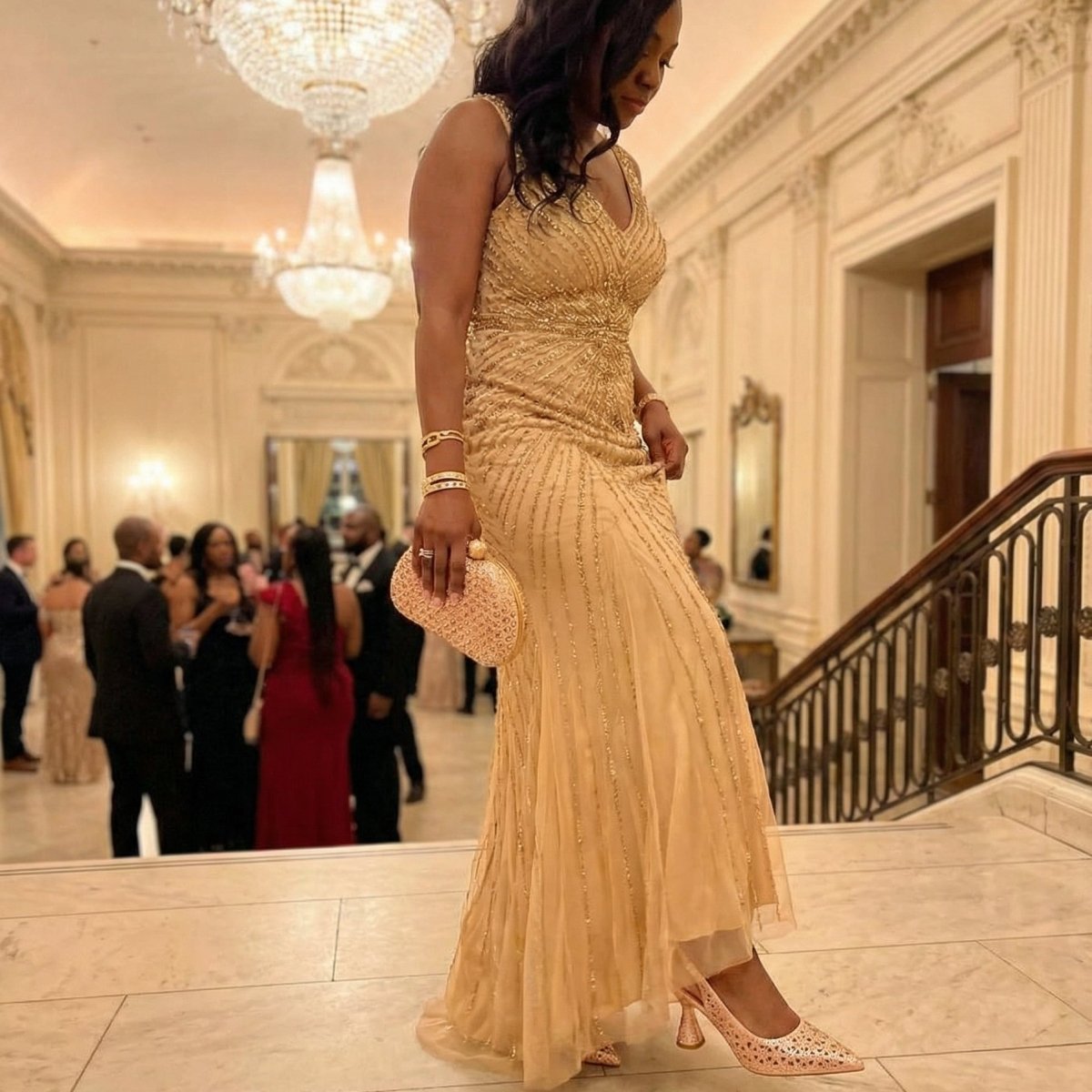 Woman wearing champagne Italian shoe and bag set elegant diamond party heels in a gold evening gown standing in an elegant indoor setting with chandeliers.