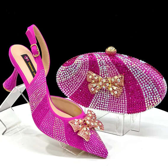 Pair of fuchsia high-heeled shoes with matching embellished clutch on a clear stand against a black background.